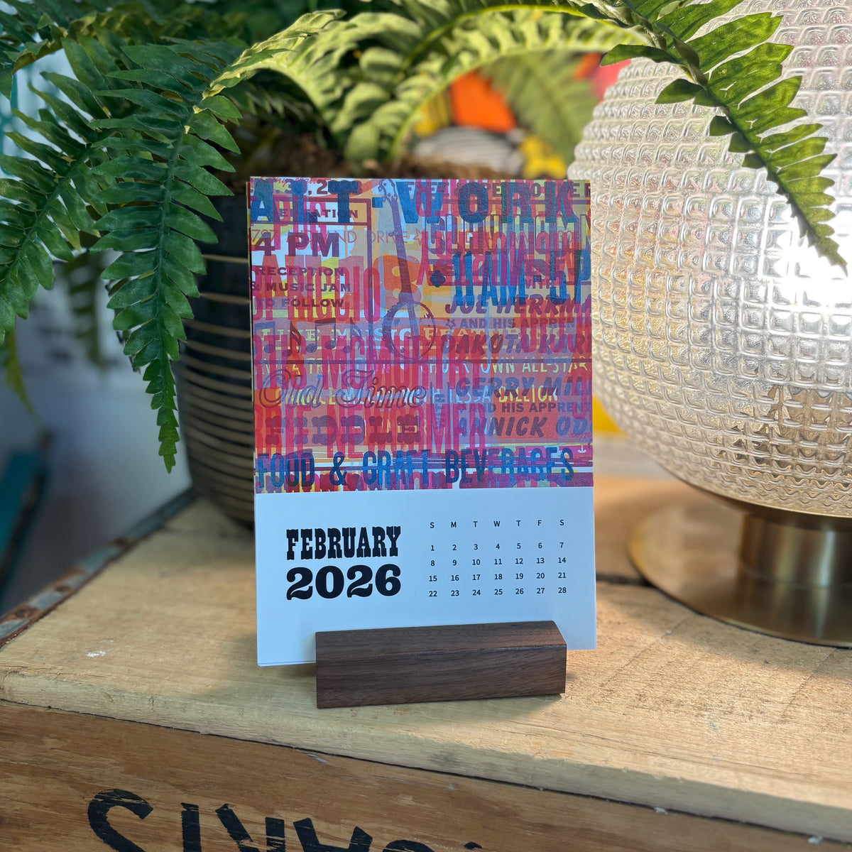 Colorful February 2026 calendar on a wooden surface with plants and a lamp in the background.